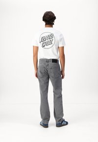 T-shirt blanche à manches courtes avec le logo "Santa Cruz" en noir, associée à un jean en denim gris et des baskets rayées bleu. Design simple, coupe décontractée.