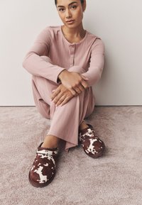 Pantofole a fantasia marrone e bianca con finitura in tessuto morbido, indossate con abbigliamento da casa a coste rosa chiaro, su una superficie di moquette soffice.