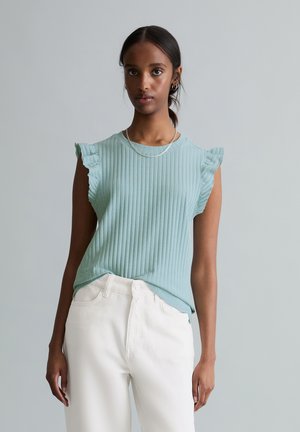 Junge Frau trägt ein hellgrünes, geripptes, ärmelloses Oberteil mit gerafften Schultern und weiße, hoch taillierte Hose, stehend vor einem grauen Hintergrund.