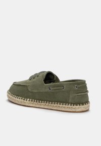 Maslinasto zelena espadrila s mekanim platnenim gornjim dijelom, zaobljenim vrhom i kontrastnom jute užadnom petom. Ima metalne oči i šivane detalje.