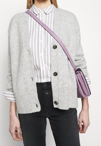 Cardigan en tricot gris clair avec une coupe décontractée, présentant des boutons foncés. Porté sur une chemise rayée blanche et grise, associé à un jean noir.