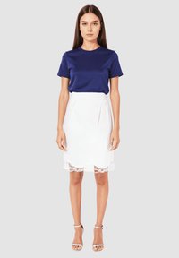 Mujer joven de pie, vistiendo una camisa azul de manga corta, una falda blanca hasta la rodilla con un dobladillo de encaje y tacones blancos con correa al tobillo, fondo neutral.