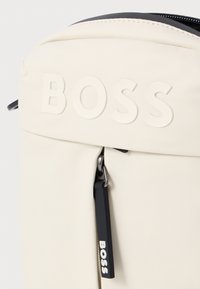 Bijela tkanina ruksak s teksturiranim završetkom, ugraviranim logotipom "BOSS" i crnim povlačnim tagom s oznakom "BOSS".