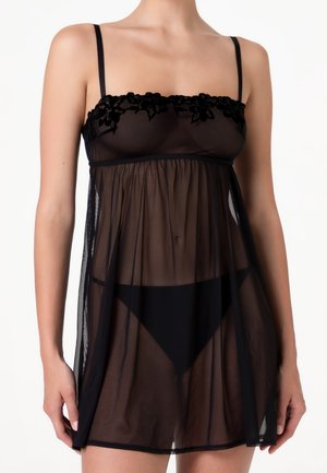 Donna che indossa un babydoll nero trasparente con ricami floreali sul busto e sottili bretelle, in piedi davanti a uno sfondo bianco.