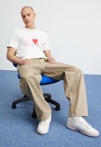 Vit t-shirt med rosa grafisk design och röda hjärtan, beige vida byxor och vita sneakers på en blå kontorsstol mot en enkel bakgrund.
