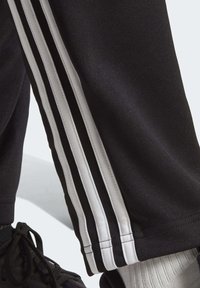 Pantalons de sport noirs avec trois bandes blanches sur le côté, fabriqués en tissu lisse avec une coupe ajustée. Baskets noires et chaussettes blanches visibles.