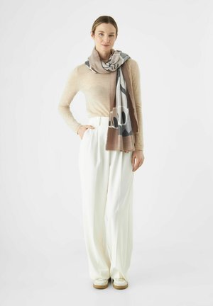 Lichte, gemusterde sjaal in beige, grijs en wit, gedrapeerd om de nek. Model draagt een crèmekleurige broek en een aansluitende, lichte trui.