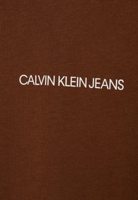 Hněd�á látka se textem „CALVIN KLEIN JEANS“ v tučných bílých písmenech, vycentrovaným a výrazným na materiálu.