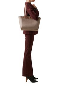 Woman in burgundy suit and black heeled boots carrying large beige textured tote bag over shoulder.