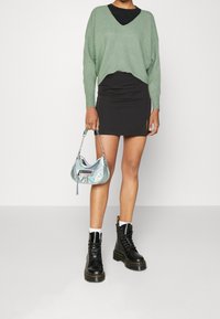 Maglione verde corto, gonna nera mini, borsa iridescente lucida e stivali combat neri con calzini bianchi. Design semplice e moderno.