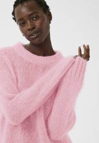 Pull rose en maille avec coupe ample, larges poignets et encolure côtelée ; texture douce et duveteuse créée par le fil.