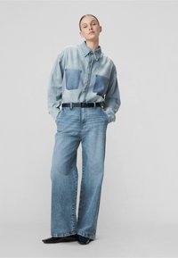 Camisa de mezclilla con dos bolsillos de parche azules, tono azul claro; combinada con jeans sueltos de color azul claro y un cinturón negro. Textura suave, zapatos pulidos.