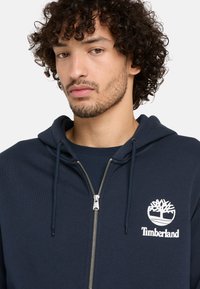 Sudadera con cremallera de color navy, hecha de una mezcla de algodón, con bolsillo tipo canguro, capucha con cordón y logo blanco de Timberland en el pecho izquierdo.