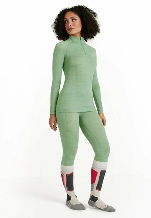 Maglia termica verde chiaro a maniche lunghe con zip a metà e leggings abbinati. Presenta motivi testurizzati. Calzini grigi e rossi con accenti distintivi.