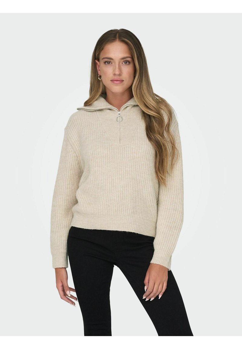 Pull en maille beige avec un col haut et une petite fermeture éclair, présentant un motif texturé et une coupe décontractée, associé à un pantalon noir.