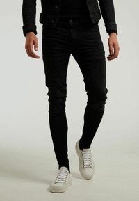 Mann trägt schlanke, schwarze Jeans mit Used-Look, schwarze Jacke und weiße Sneaker, geht vor einem einfarbigen hellen Hintergrund.