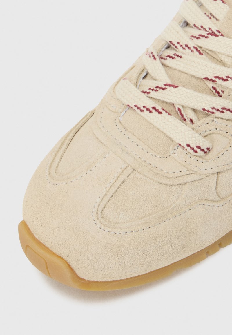 Sneaker in suede beige con superficie texturizzata, lacci a contrasto rossi e crema, e suola in gomma per una migliore aderenza. Presenta dettagli cuciti.