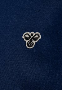 Marinblått stickat tyg har en liten broderad bi-logotyp i vitt och svart, vilket visar på intrikata detaljer och textur.