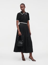 Femme portant un polo noir à manches courtes avec une bordure blanche, une jupe midi noire avec de petites décorations, un sac à main noir et des sandales noires à talons.