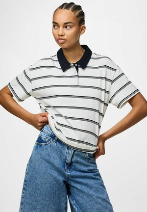 Mladá osoba s vrkočmi, oblečená v bielej krátkorukávovej polokošeli s čiernymi pruhmi a modrých rifle s vysokým pásom, pózujúca s rukami na bokoch.