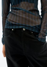 Ein langärmliges, transparentes Oberteil in dunkler Farbe mit einem gestreiften Muster, kombiniert mit hoch taillierten schwarzen Jeans mit silbernen Knopfakzenten.