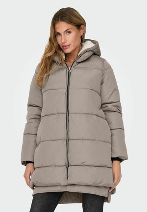Femme portant une veste d'hiver rembourrée taupe avec capuche et fermeture éclair à l'avant, se tenant avec les mains relâchées sur les côtés contre un fond uni.