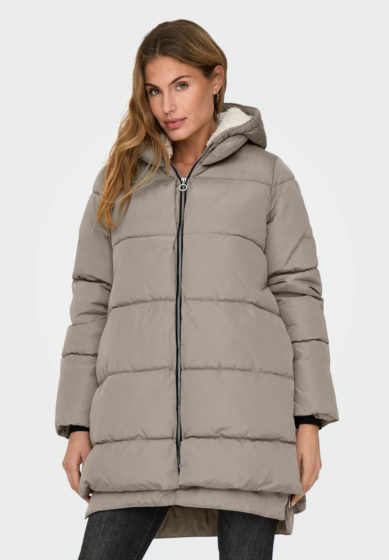 Femme portant une veste d'hiver rembourrée taupe avec capuche et fermeture éclair à l'avant, se tenant avec les mains relâchées sur les côtés contre un fond uni.