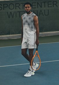 Conjunto atlético branco com uma parte superior de tank top estampada e calções, combinado com ténis brancos e uma raquete de ténis laranja num court.