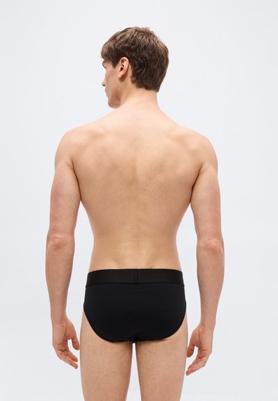 Homme aux cheveux bruns courts vu de dos, portant un slip noir sur un fond gris clair uni.