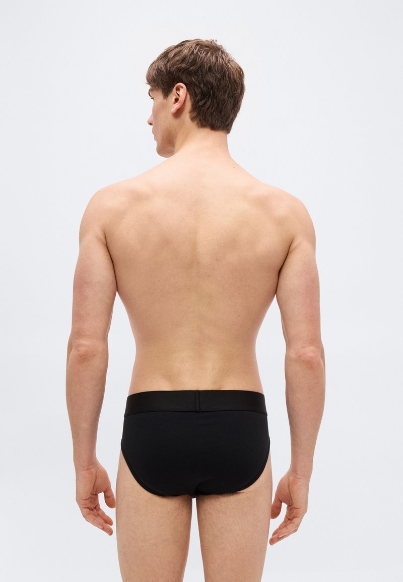 Man with short brown hair viewed from behind, wearing black briefs against a plain light gray background.