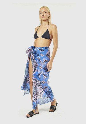 Donna che indossa un top bikini nero, un sarong con motivi blu e viola legato in vita e sandali neri, in piedi su sfondo bianco.