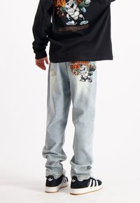 Lichtblauwe denim jeans met een cartoonafbeelding van een personage op het achterzakje. Gecombineerd met een zwart shirt met lange mouwen en zwarte sneakers.