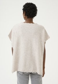 Licht beige oversized gebreide poncho met golvende zoom en korte mouwen, gemaakt van zachte stof, gedragen over grijze jeans.
