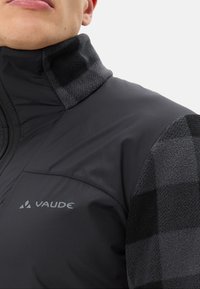 Giacca imbottita nera e grigia con colletto in pile, caratterizzata da un logo sul petto e un motivo a scacchi sulla spalla.