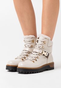 Bottines en cuir beige clair avec une bordure en fausse fourrure, des œillets dorés et une boucle. Semelle en caoutchouc noire avec accent en bois. Design à lacets.