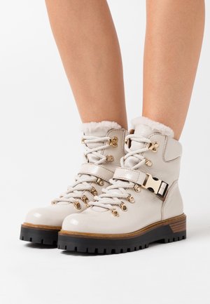 Botas de tobillo de cuero beige claro con borde de pelo sintético, ojales dorados y un hebilla. Suela de goma negra con acento de madera. Diseño con cordones.