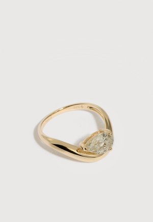 Anillo de oro con un diamante en forma de pera colocado de forma asimétrica sobre una banda pulida y retorcida, sobre un fondo claro.