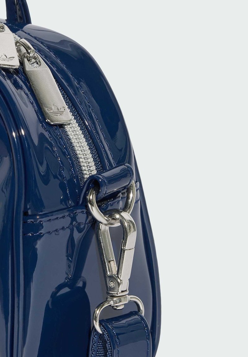 Ecke einer glänzenden, marineblauen Tasche mit silbernen Reißverschlussanhängern, die Adidas-Logos zeigen, und einem silbernen Drehverschluss, der an einem Riemen befestigt ist.