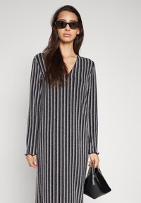 Robe longue rayée avec des lignes verticales métalliques argentées et noires, décolleté en V et manches longues. Le mannequin tient un petit sac à main noir.