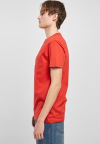Rotes Baumwoll-T-Shirt mit kurzen Ärmeln, Rundhalsausschnitt, lockerer Passform und glatter Textur. Getragen mit blauen Jeans, von der Seite betrachtet.