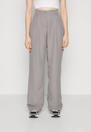Pantalons larges gris en tissu lisse, avec une fermeture à bouton, des plis doubles, et reposant au-dessus de baskets noires et blanches.