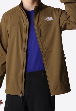 Personne portant une veste North Face marron zippée par-dessus une chemise bleue et un pantalon noir, posant avec un bras levé.