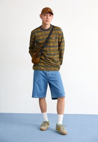 Long-sleeve striped shirt in green and orange, denim shorts, tan cap, and gray sneakers with yellow and green accents. Small brown bag crossbody.