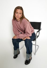 Ružový top s dlhými rukávmi a naťahovacími rukávmi, denimové džínsy so šedými manžetami a čierne mokasíny. Proti jednoduchému bielej pozadiu.