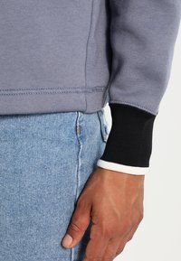 Grå sweatshirt med svarta och vita ribbade mudd, parat med ljusblå denimbyxor. Har texturerad tyg och synliga sömmar.