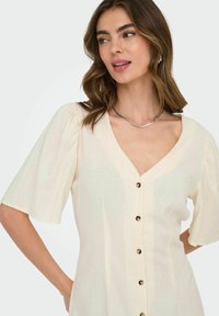 Abito color crema con maniche a sbuffo corte, scollatura a V e bottoni decorativi lungo il davanti. Tessuto texturizzato con un aspetto leggero e fluido.