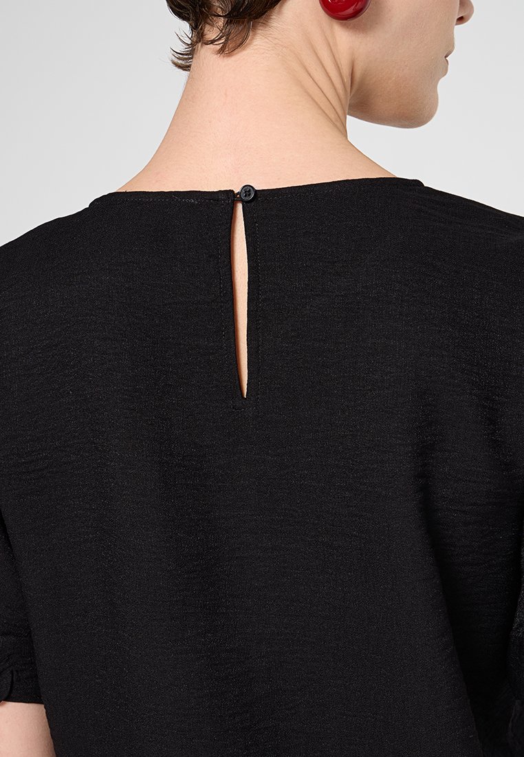 Schwarzes Oberteil aus strukturiertem Stoff, mit rundem Ausschnitt und einer kleinen Schlüssellochöffnung hinten, die mit einem einzelnen Knopf gesichert ist.
