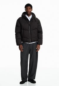 Schwarze gesteppte Jacke mit Kapuze, strukturiertem Stoff, Reißverschluss vorne und Seitentaschen. Kombiniert mit grauen Nadelstreifenhosen und schwarzem Schuhwerk.