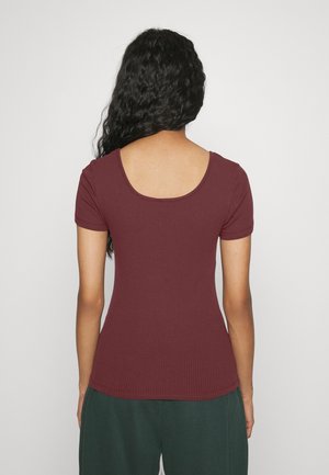 Mujer con cabello largo y rizado, vistiendo una camiseta de manga corta color burdeos con textura acanalada y espalda en forma de escote redondo, y pantalones verde oscuro, de espaldas a la cámara.