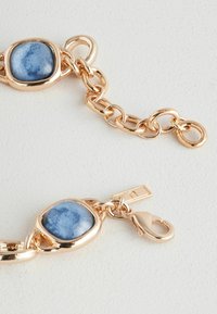 Bracelet en chaîne dorée avec une accents en pierre bleue. Le fermoir est de style homard avec une étiquette plate attachée. Fini métallique lisse et brillant.